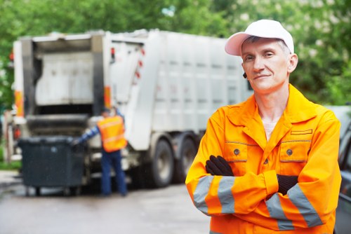 A professional waste management worker from Office Clearance Palmers Green wearing an orange high-visibility jacket with reflective stripes, a white cap, and black gloves, standing with arms crossed in front of a large white rubbish collection truck parked on a paved area. In the background, another worker in similar attire is loading or unloading waste materials into the truck, with a green, leafy environment suggesting an outdoor setting near Palmers Green. The scene depicts a typical rubbish removal operation involving the collection of waste from commercial or residential premises, with the truck's rear open, revealing the compaction mechanism inside. The image highlights the practical aspects of rubbish clearance services, including safety clothing, loading equipment, and environmental surroundings, aligning with the services offered by Office Clearance Palmers Green for efficient waste collection and disposal within the local area.