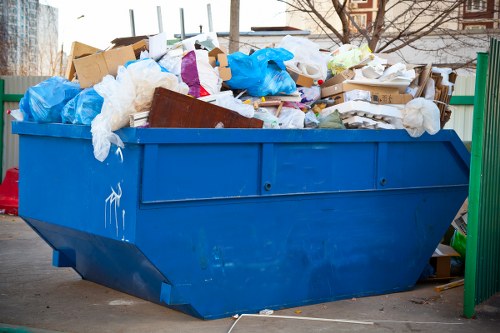 A large blue metal skip positioned outdoors on a paved surface, overflowing with mixed waste materials including brown cardboard boxes, white plastic packaging, blue plastic bags, and various paper and plastic debris. The skip is situated near a green metal fence, with a background of leafless trees and a building, suggesting a winter or early spring setting. The waste appears haphazardly piled, with some materials spilling over the edges, indicating recent rubbish removal activity typical of commercial or domestic clearance services in Palmers Green, North London. The overall scene reflects a typical rubbish disposal scenario handled by companies like Office Clearance Palmers Green, focused on clearing unwanted waste efficiently from properties in the vicinity.