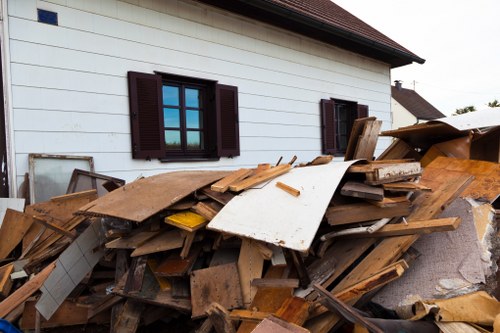 A large pile of discarded wooden planks, broken furniture, and miscellaneous construction debris is stacked against the side of a white residential building with dark brown window shutters, located on a paved surface. The timber varies in size, color, and texture, with some pieces appearing weathered and others smoother. Some panels and broken boards are leaning randomly, creating a cluttered appearance. To the right of the pile, there is a cardboard box partly filled with more waste material. The building's exterior walls are smooth, painted white, and feature three visible windows with matching dark brown louvered shutters. In the background, other rooftops and trees can be seen, indicating a suburban setting. The scene suggests a site awaiting rubbish removal or clearance, consistent with services provided by Office Clearance Palmers Green in the local area. The lighting appears natural, possibly on an overcast day, contributing to the neutral tone of the image.