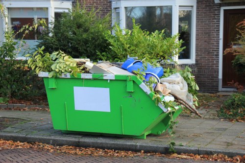 A large, bright green skip filled with various types of waste, including green leafy branches, cardboard boxes, plastic packaging, and miscellaneous rubbish, positioned on a paved driveway in front of a residential brick building with white-framed windows. The skip is placed on the pavement adjacent to a curb, with some surrounding greenery and bushes visible behind it. The waste appears to be in the process of collection or removal, consistent with services offered by Office Clearance Palmers Green for local rubbish disposal needs in the area near Palmers Green, UK.