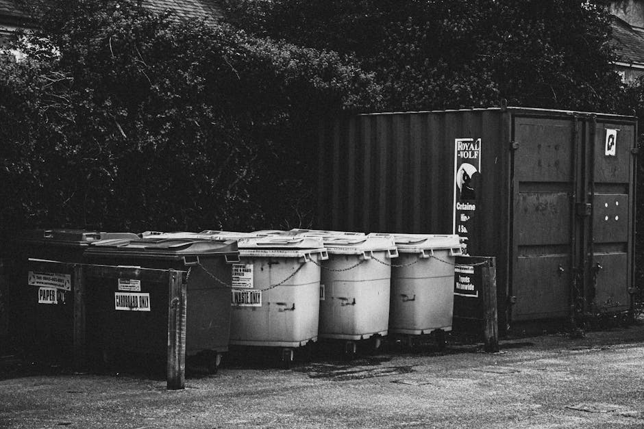 A row of five large plastic wheelie bins with closed lids, positioned on a paved surface next to a metal container. The bins are primarily light in colour, with some displaying labels indicating their