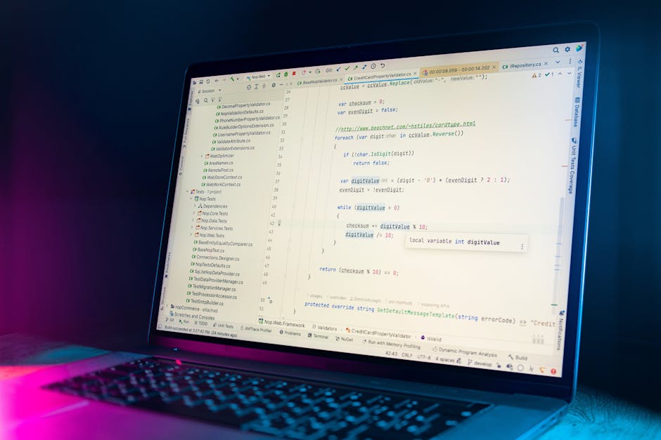 A close-up view of a person's hands typing on a laptop keyboard placed on a dark, reflective table. The laptop screen displays lines of code with a dark background and multicoloured syntax highlightin